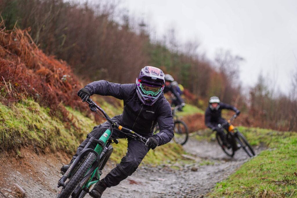 Riders wearing winter MTB gear essentials including waterproof jackets, gloves, and goggles on a wet, muddy trail — essential cold-weather mountain bike kit in action.